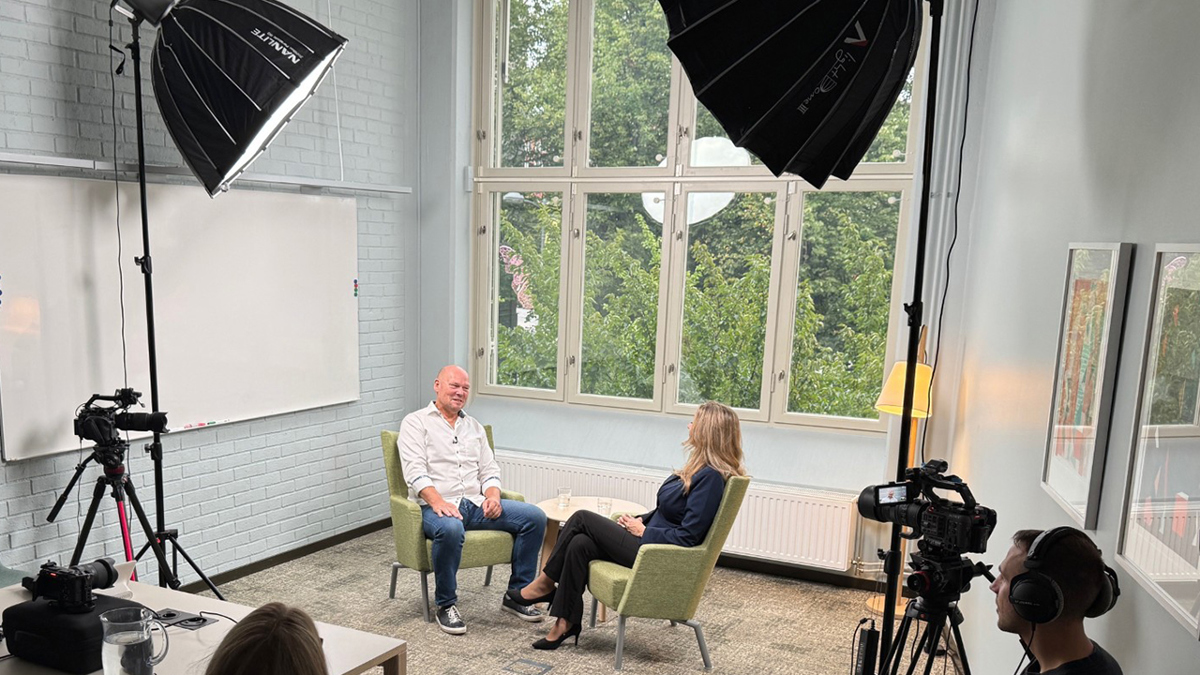 Två personer sitter i ett rum med stort fönster. Utanför syns gröna träd. Inne i rummet finns stora lampor och filmkameror. En filmare med hörlurar sitter i förgrunden.