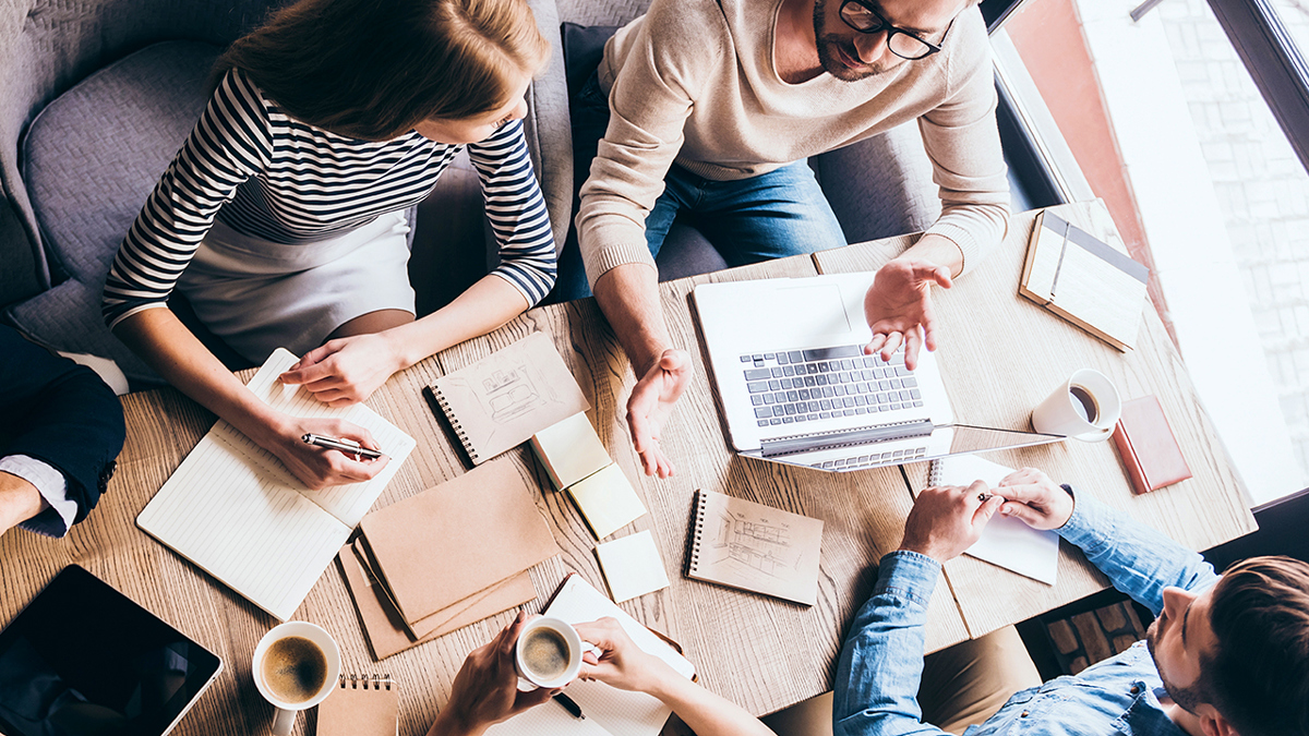 Vy från ovan: människor runt ett bord fyllt med anteckningsblock, kaffekoppar och en dator