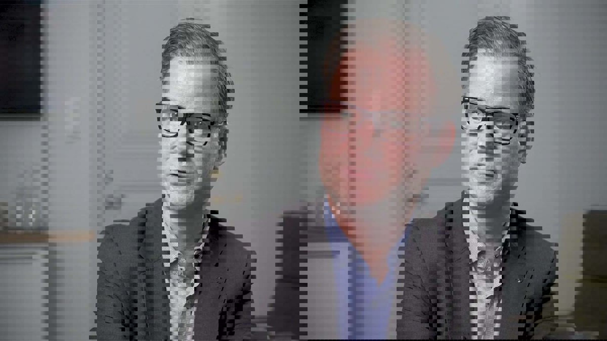 Person införd mörkgrå kavaj, blå skjorta och svartbågade glasögon.