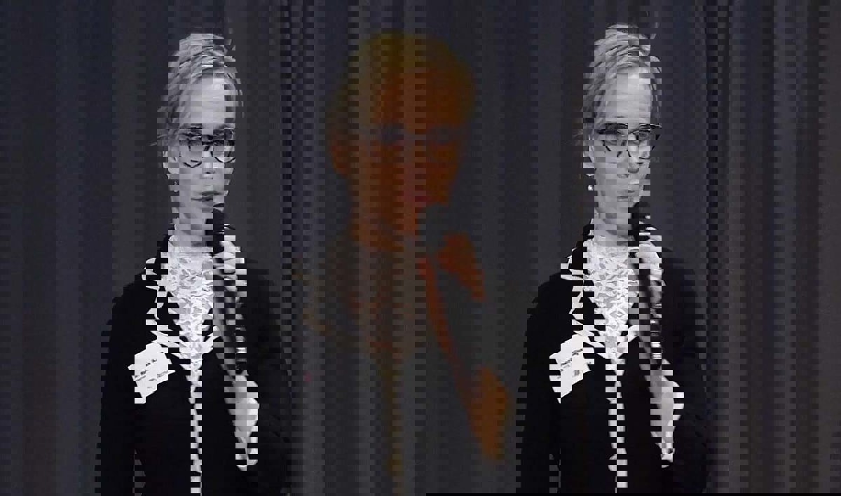 Person i föreläsningssituation hållandes en mikrofon iförd svart kavaj och vit blus.