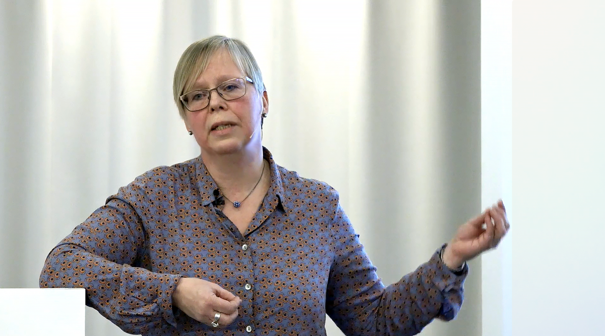 Kvinna vid podium framför vit gardin.