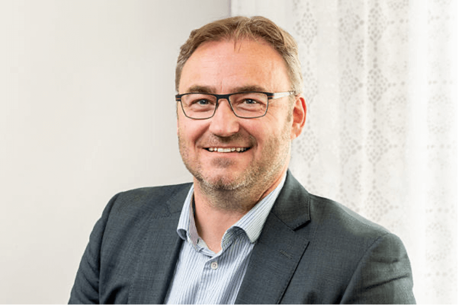 Person iförd glasögon, grå kostym och blårandig skjorta.