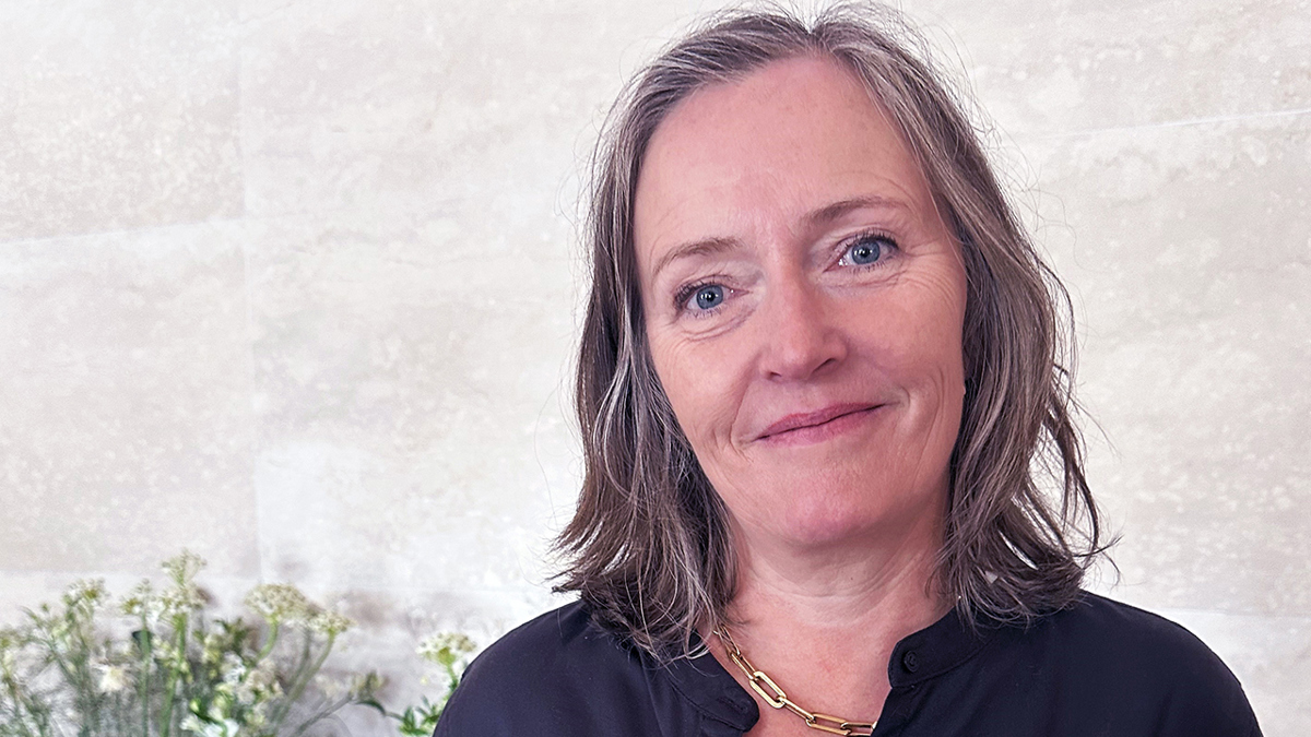 Person iförd svart blus och halsband, stående framför en bukett blommor.