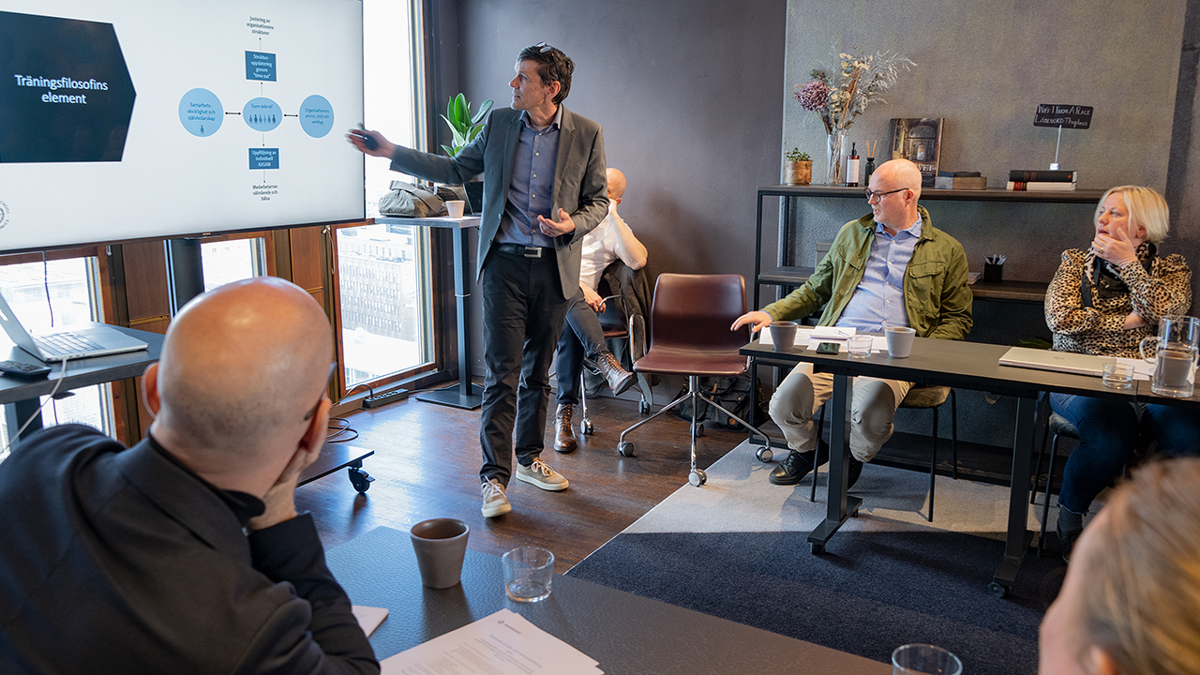 En föreläsare står framför en ppt-presentation medan flera andra tittar och lyssnar på honom.