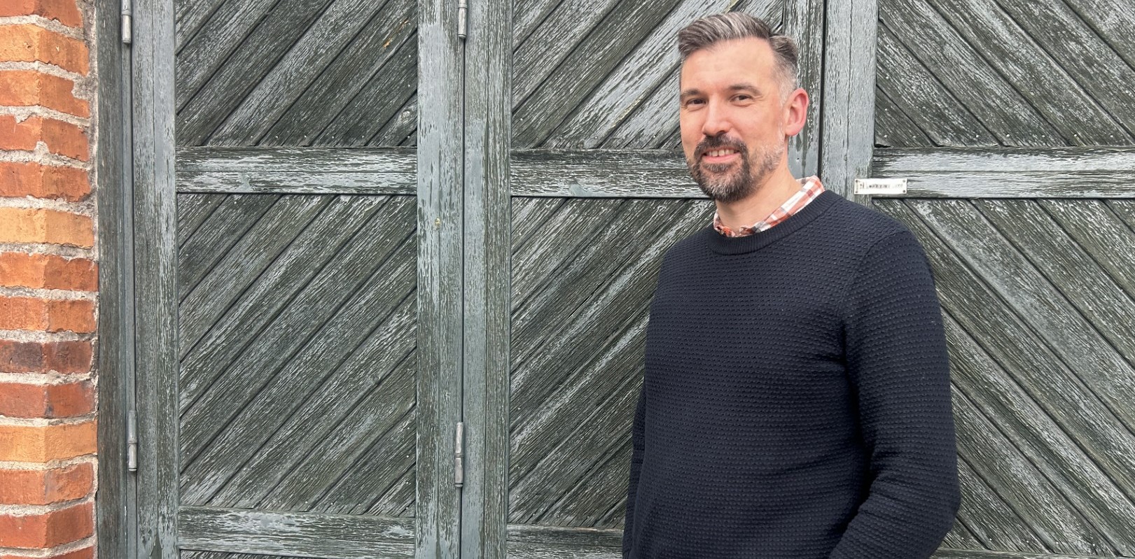 Person står vid en gammal grånad träport. En tegelvägg syns till vänster. Personen syns i halvkropp och ler mot kameran.