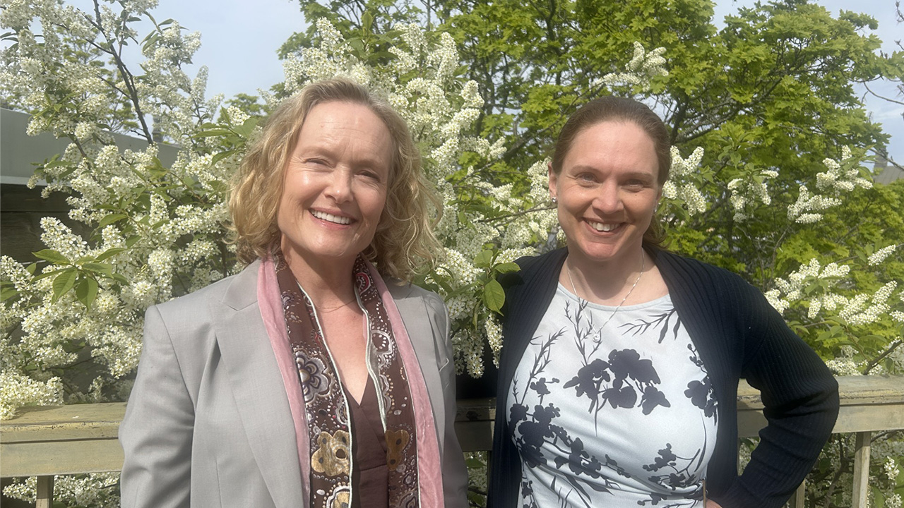 Två personer står utomhus framför en blommande hägg. De ler och tittar in i kameran.