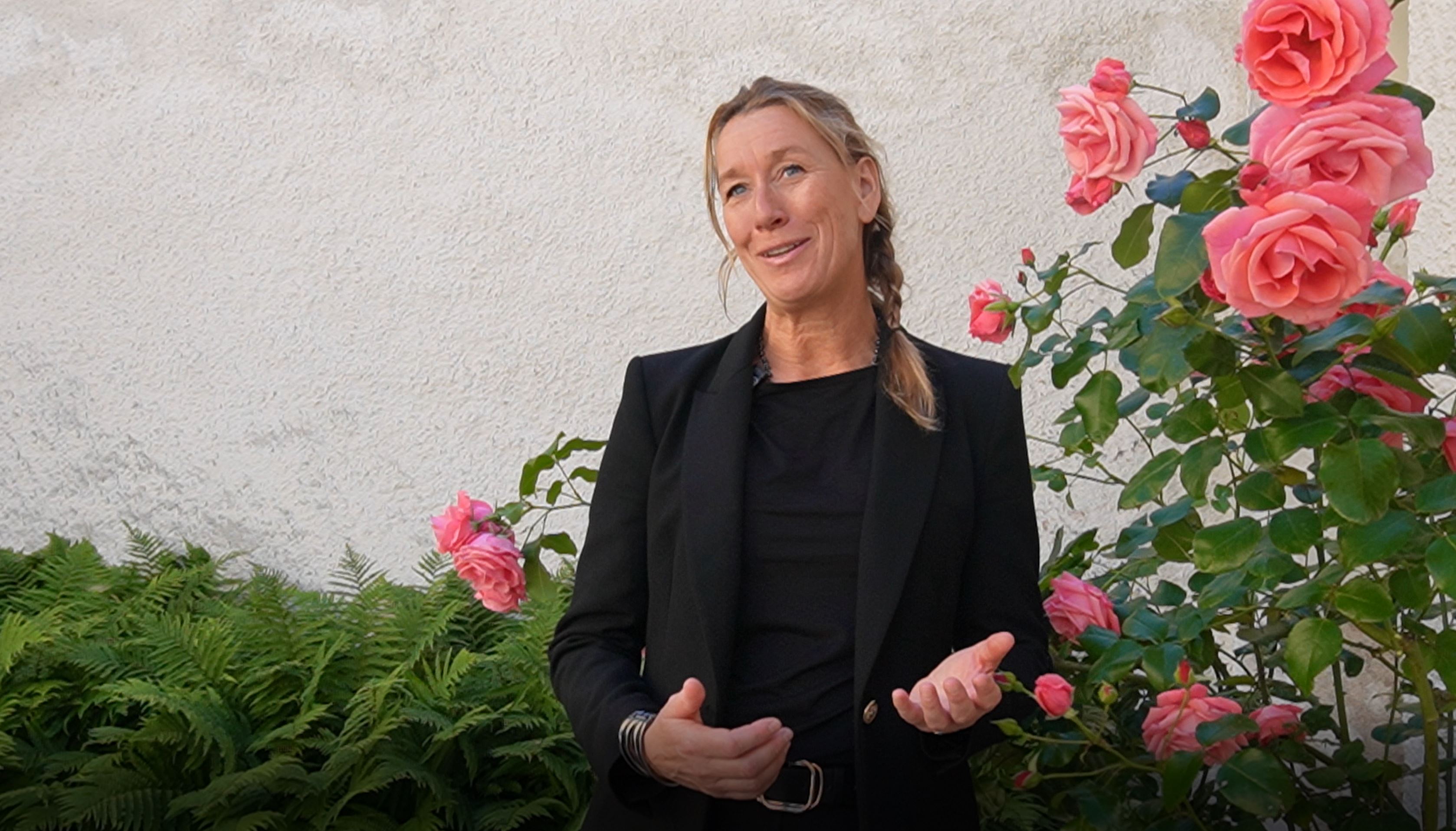 Kvinna står och pratar in i kameran utomhus med rosor bredvid sig.