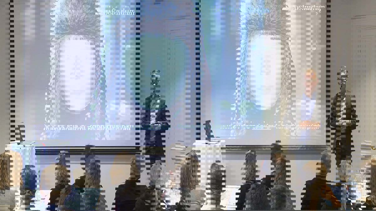 Person i föreläsarsituation stående bredvid en powerpointbild med en bild av en robot med pälsmössa.