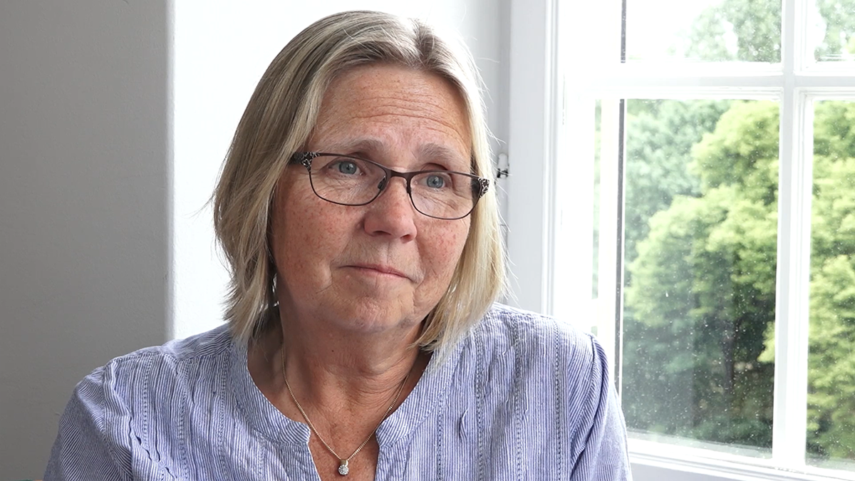 Perons iförd randig blus sittandes vid ett fönster där man ser stora lummiga träd.