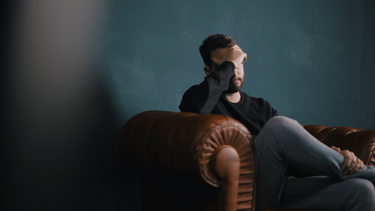 Ledsen person sitter i fåtölj med handen mot pannan i en uppgiven gest.