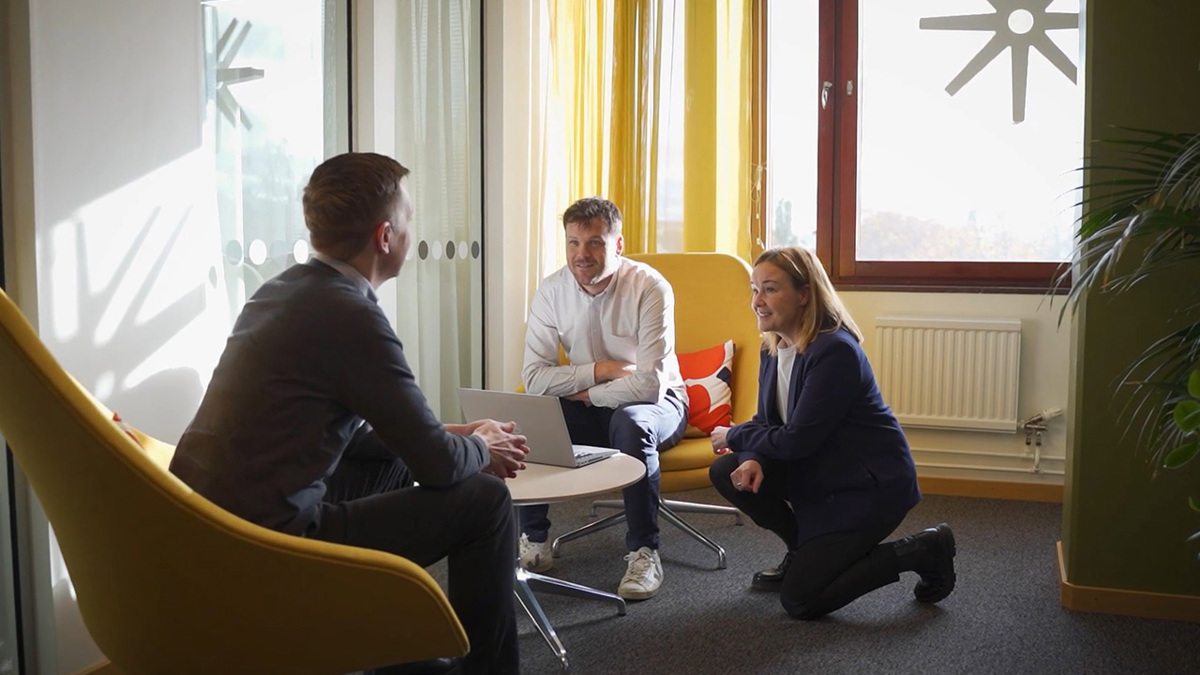 Två personer sitter i fåtöljer i en kontorskorridor och pratar. En tredje person står på knä på golvet och lyssnar leende in samtalet.