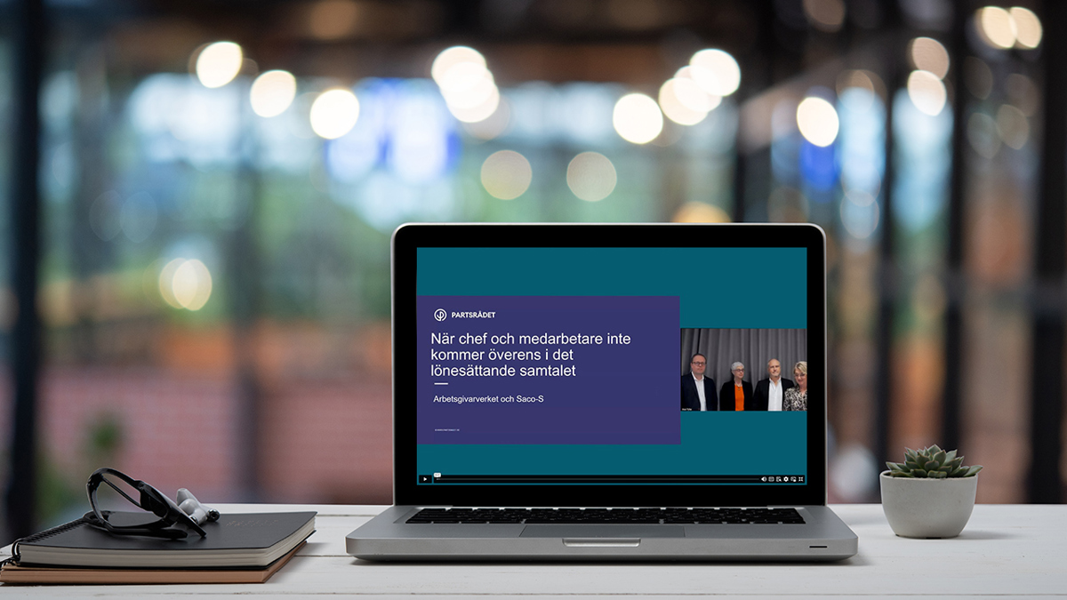 Bärbar dator står på ett bord i ett kontor. På skärmen syns ett webbinarium med fyra personer och en presentation.