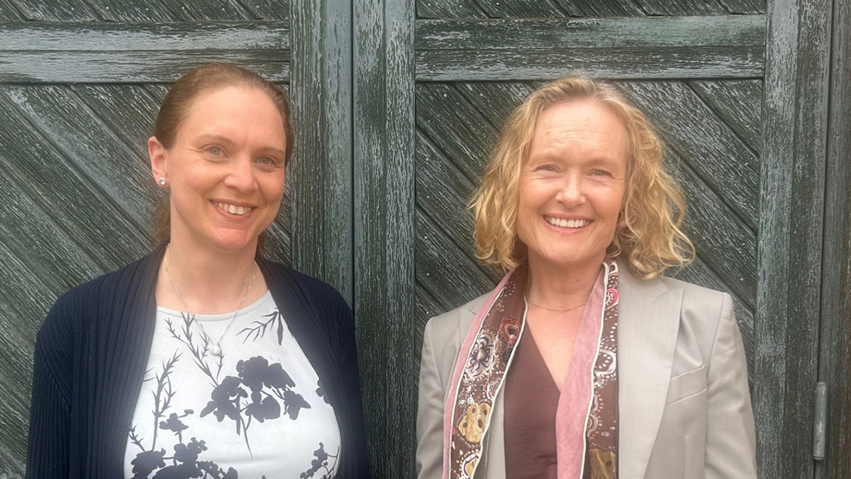 Två personer står framför en gammal grånad trädörr. De tittar in i kameran och ler.