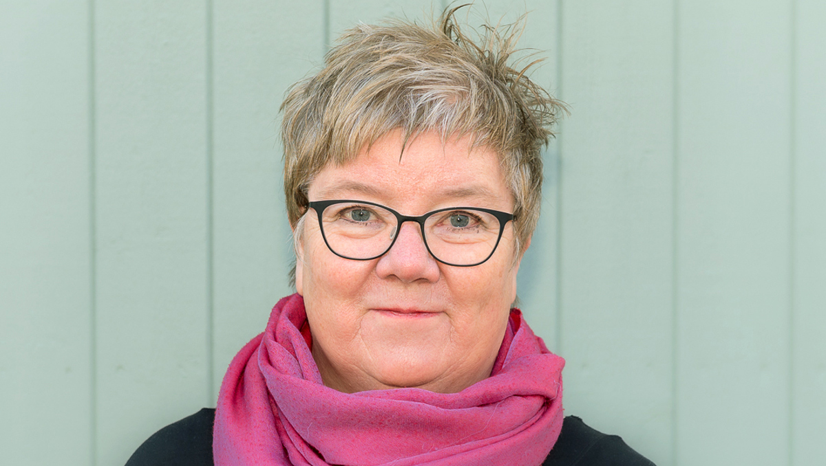 Person med kort hår, glasögon och rosa sjal, stående framför en vit vägg.