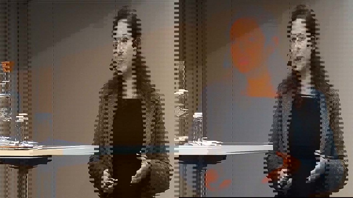 Person i seminariesituation stående bakom pulpet med vatten i en glasflaska och ett dricksglas.