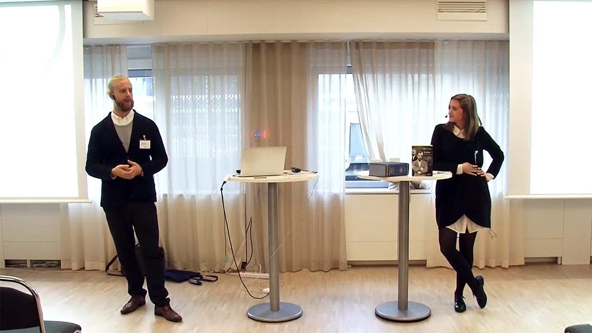Två föreläsare stående framför åhörare.
