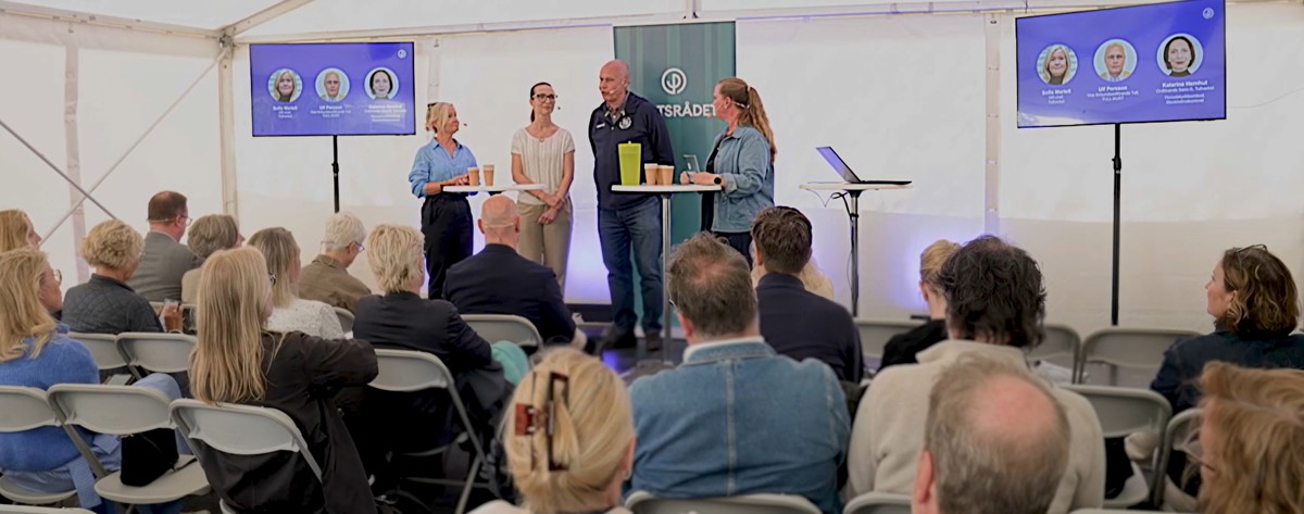 Tre personer står på en scen i ett tält och pratar med en fjärde som är moderator. Det är många i publiken.