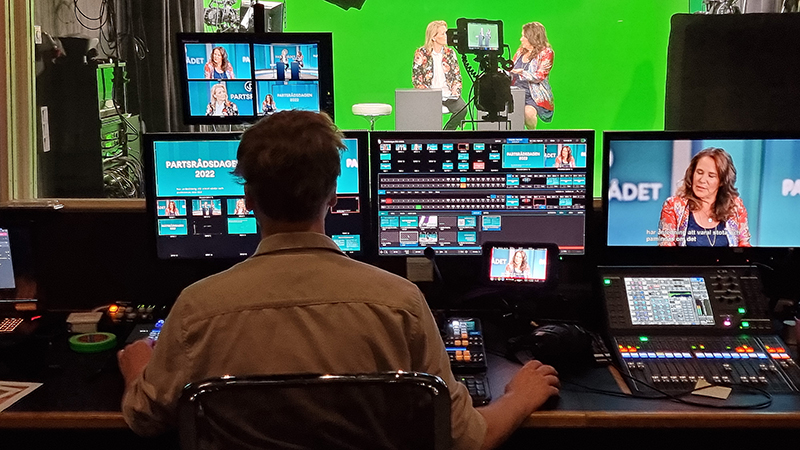 Person framför skärmar i studions kontrollrum. Genom ett fönster ser man en kamera och två personer i studion.