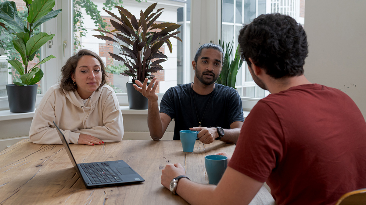 Tre personer runt ett bord med en dator i mitten och två kaffemuggar.