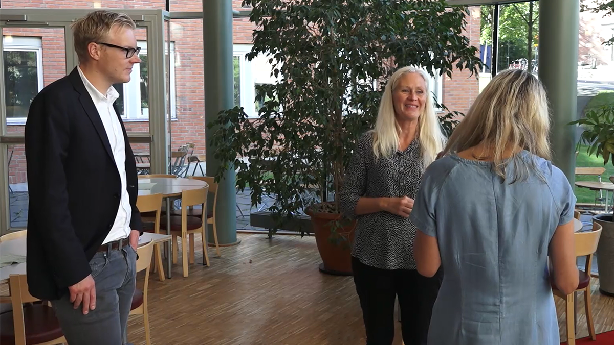 Tre personer pratar stående tillsammans i något som skulle kunna vara en lunchmatsal.
