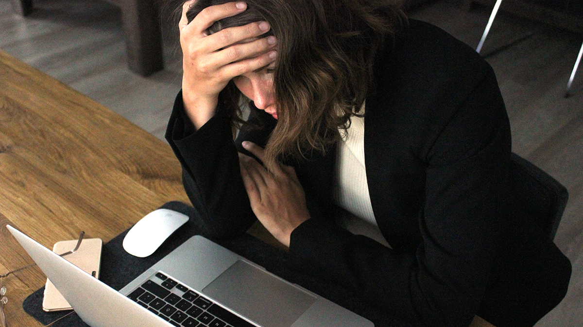 Person sitter framför dator och ser stressad ut med handen mot pannan i en uppgiven gest.