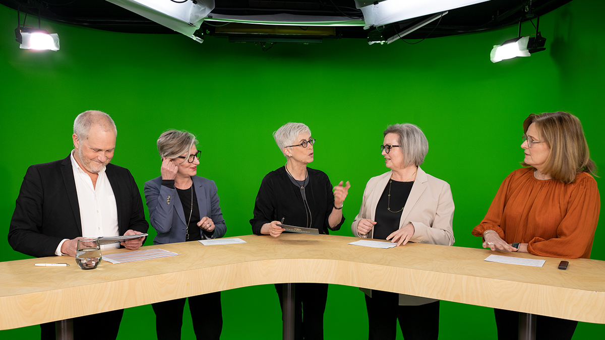 Fem personer stående vid ett högt bord under sändning.